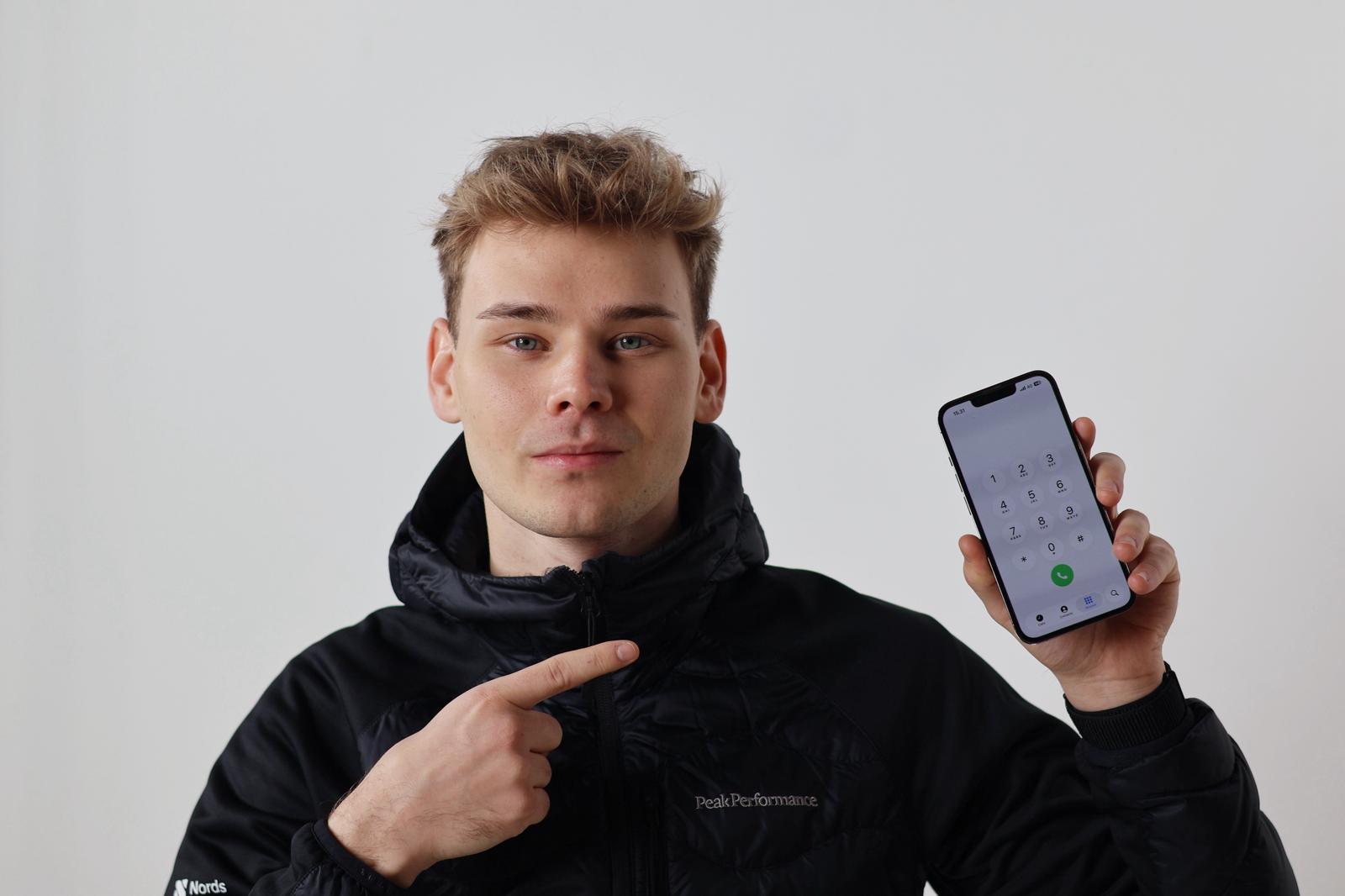 Nuori mies mustassa takissa näyttää ja osoittaa puhelintaan, jossa on näppäimistön numeronäyttö avoinna.