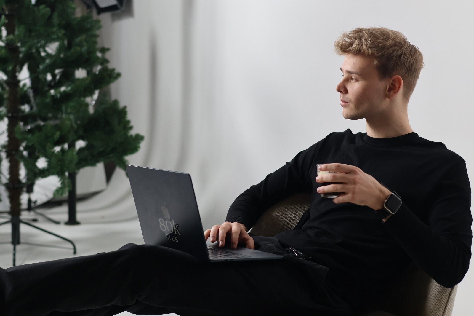 Nuori mies istuu nojatuolissa kädessään lasillinen ja sylissään kannettava tietokone, taustalla koristeltu joulukuusi.