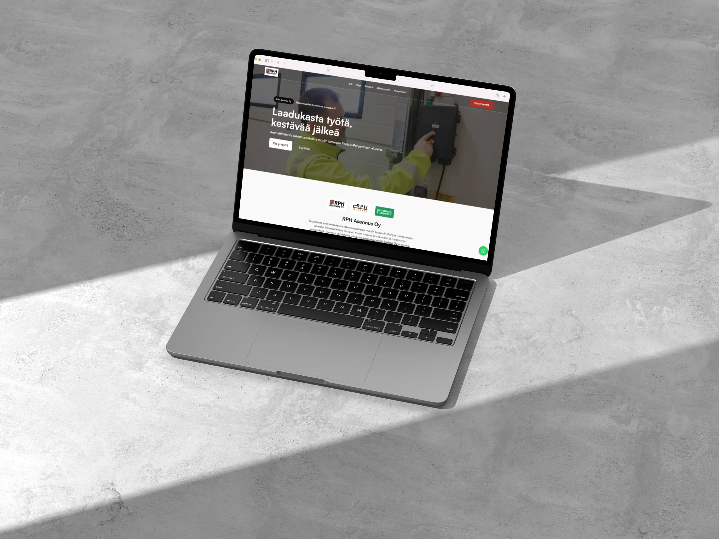 Hopeanvärinen kannettava tietokone harmaalla betonipinnalla, näytöllä RPH Asennus Oy:n verkkosivusto ja auringonvalon luoma varjo.