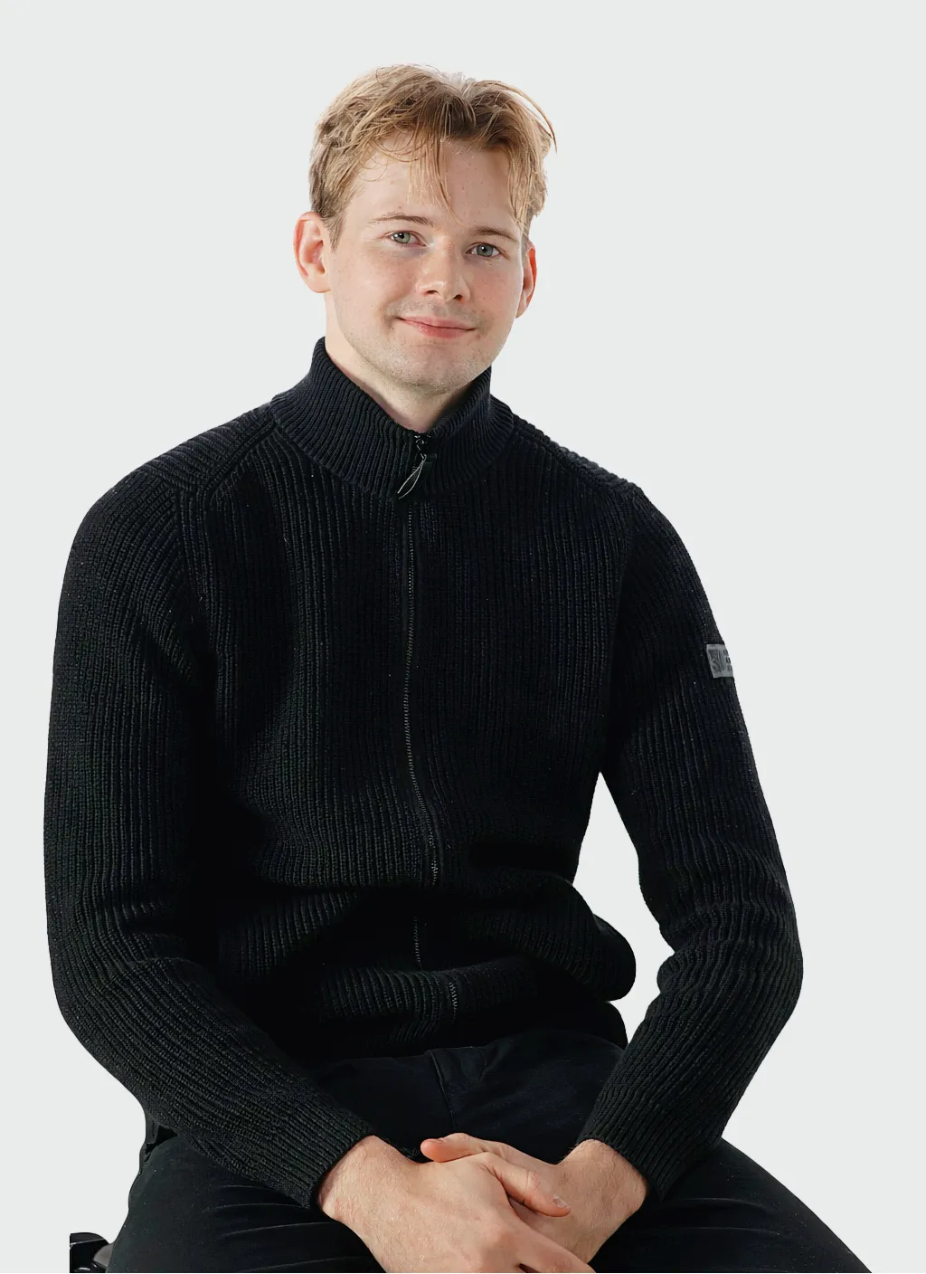 Muotokuva vaaleahiuksisesta miehestä mustassa vetoketjullisessa neuletakissa, istumassa valkoista taustaa vasten.