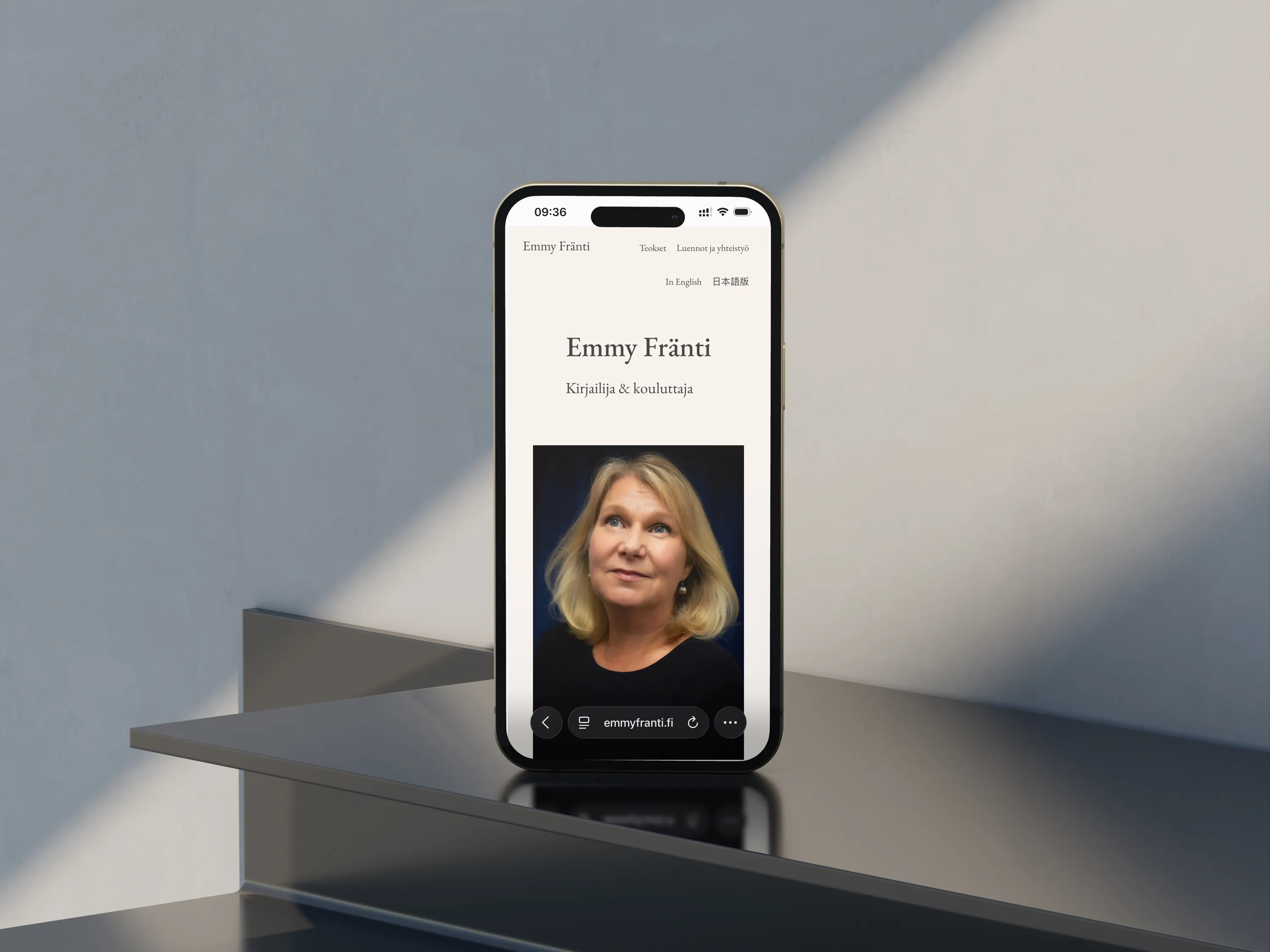 Puhelin Emmy Fräntin verkkosivulla, jossa näkyy hänen kuvansa ja tekstiä. Puhelin on sijoitettu harmaalle tasolle, ja taustalla on vaalea seinä.