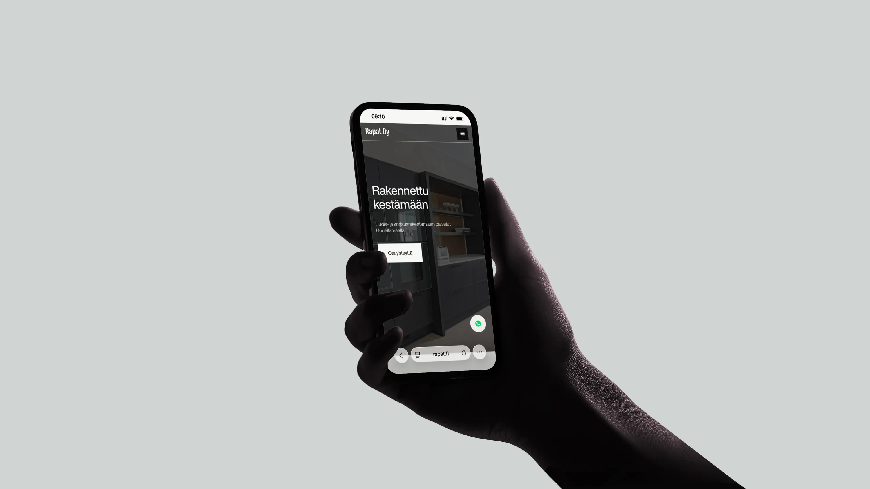 Tumma käsi pitelee älypuhelinta, jossa näkyy Rapat Oy:n verkkosivu, jossa on tekstiä ja sisustuskuva.