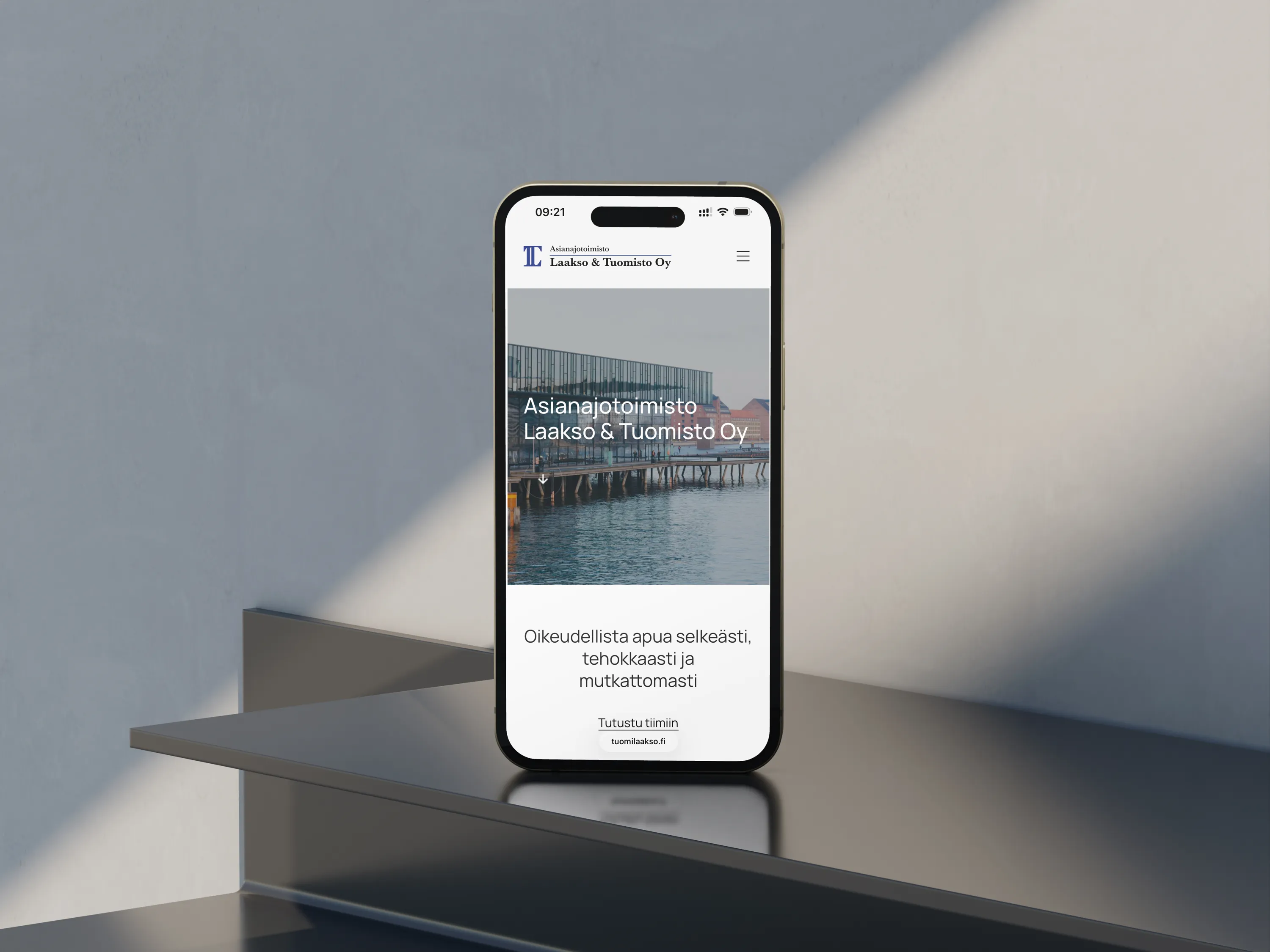 Kuva esittää älypuhelinta, jossa näkyy Asianajotoimisto Laakso & Tuomisto Oy:n verkkosivu. Puhelin on sijoitettu modernille, harmaalle tasolle, ja taustalla on vaalea seinä. Valo luo pehmeitä varjoja.